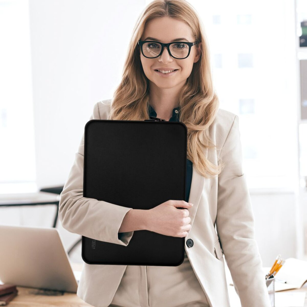 Een vrouw met bril en blazer houdt een zwarte laptoptas vast en glimlacht. Op de achtergrond een bureau en kantoorbenodigdheden.