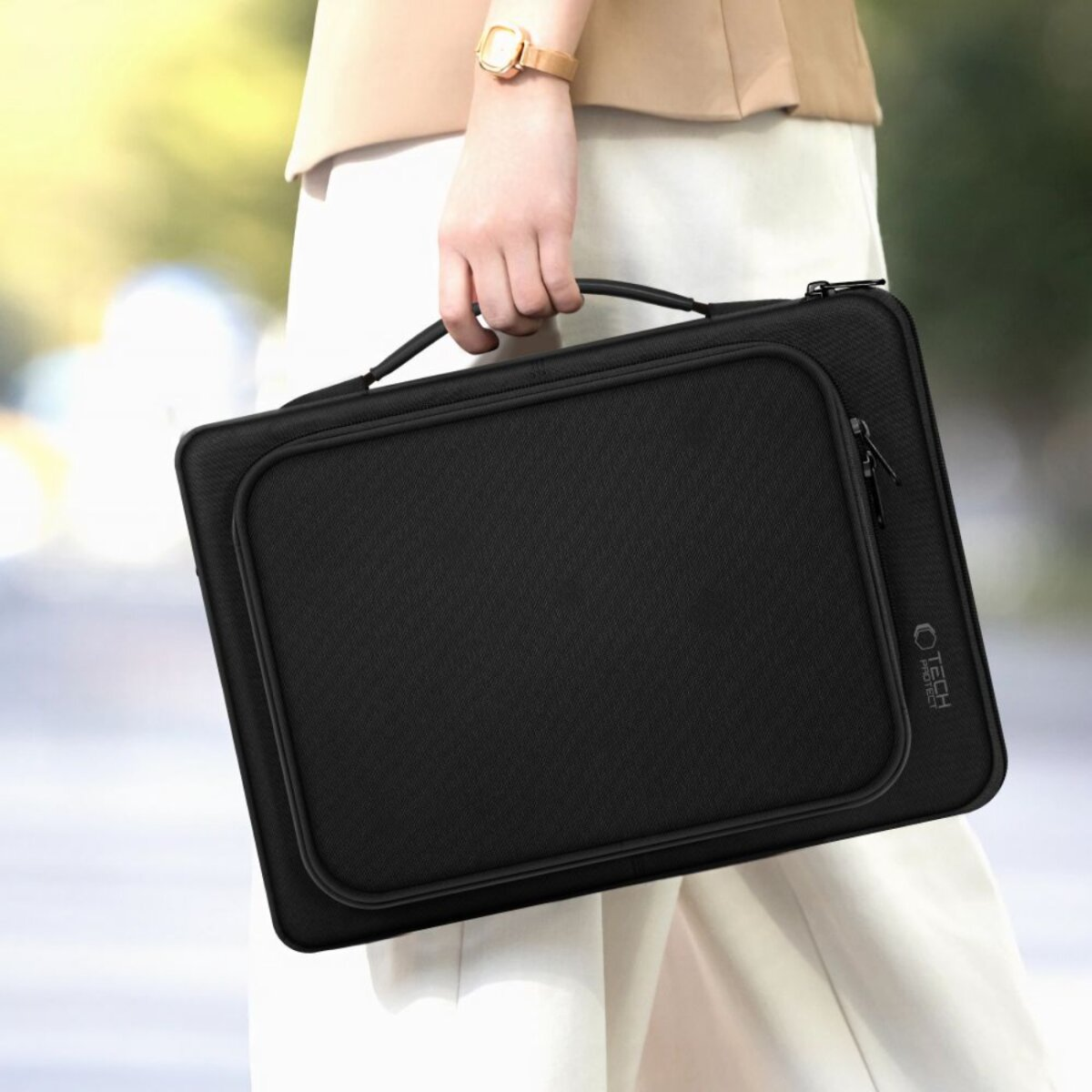 Een persoon houdt een zwarte laptoptas met een handvat vast. De persoon draagt een gouden horloge en een witte broek. De achtergrond is onscherp.