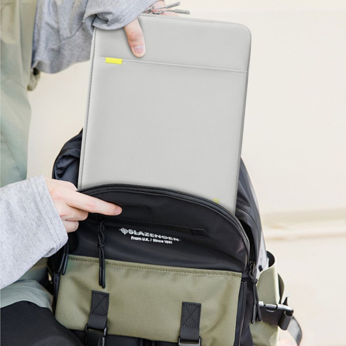 Een persoon plaatst een grijze laptoptas in een zwarte en groene rugzak. De tas heeft een gele accentkleur en een rits.