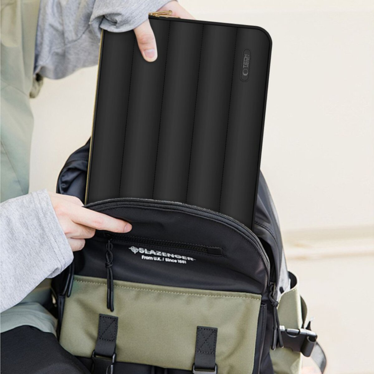 Een persoon plaatst een zwarte laptoptas in een zwarte en olijfgroene rugzak. De tas heeft een gewatteerd ontwerp en een gouden rits.
