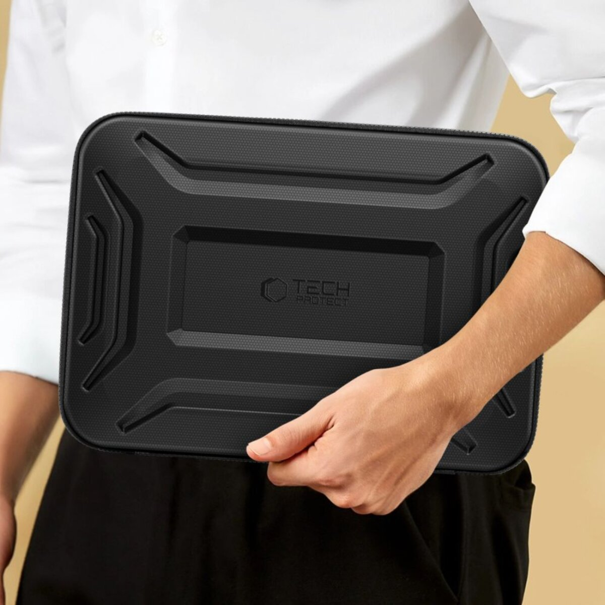 Een persoon houdt een zwarte TECH PROTECT laptophoes vast. De hoes is rechthoekig met een gestructureerd oppervlak. De persoon draagt een wit overhemd.