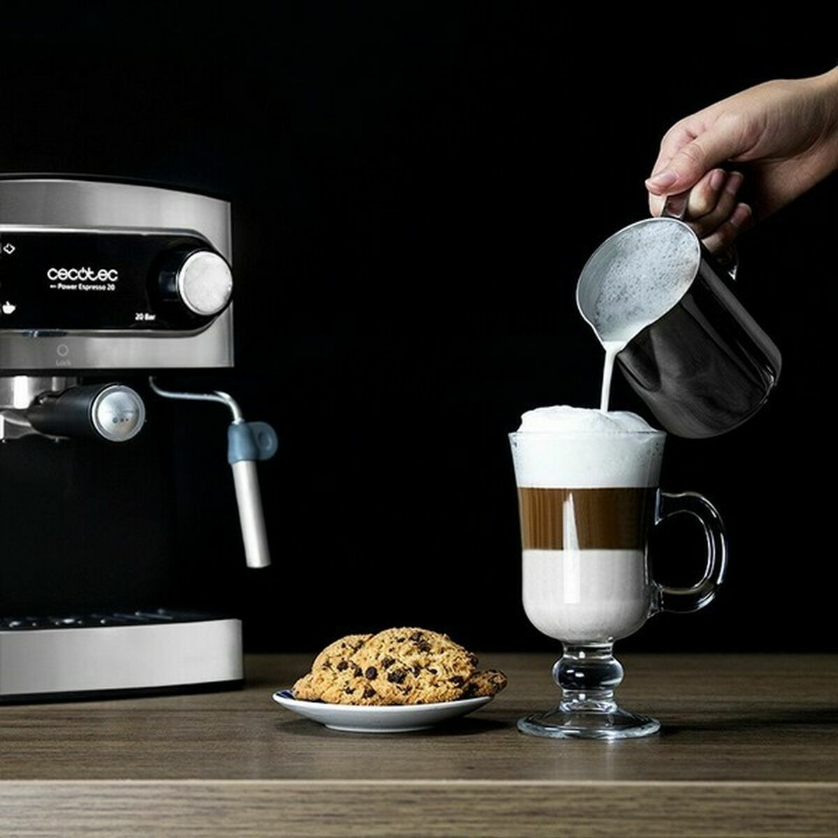 Una mano versa il latte in una bevanda al caffè a strati. Una macchina per il caffè, biscotti e un piattino sono su un tavolo di legno.