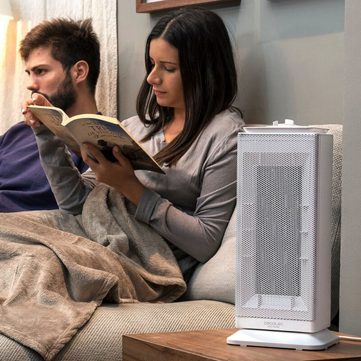 Para czyta książkę na kanapie, obok kobiety stoi biały grzejnik. Mają na sobie koc.