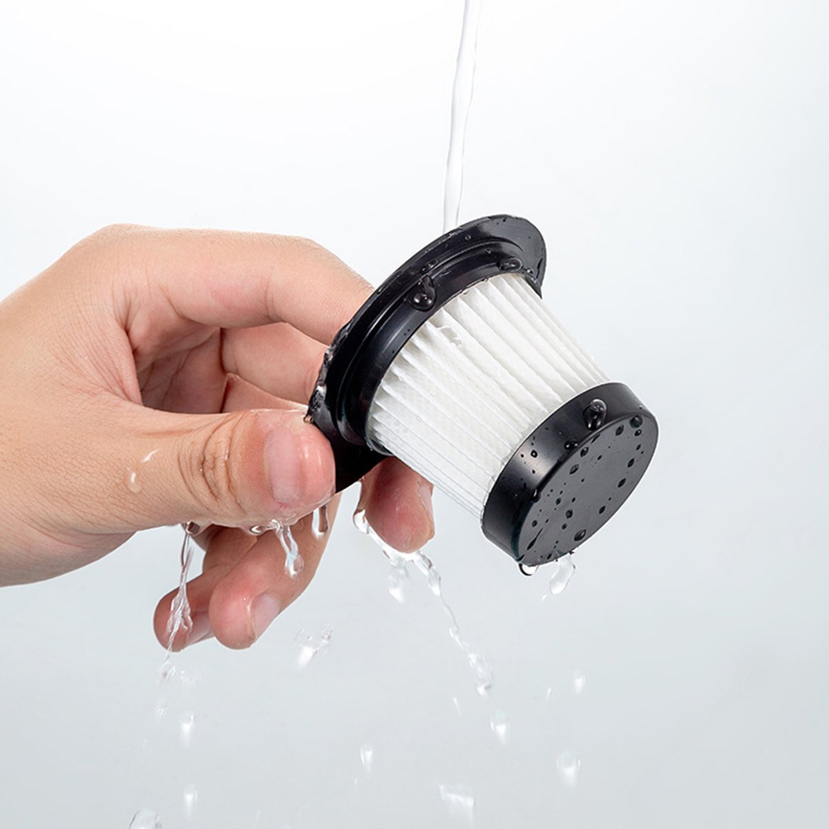 Una mano sostiene un filtro de aspiradora que se enjuaga con agua. El filtro es negro y blanco, y el agua fluye desde arriba.