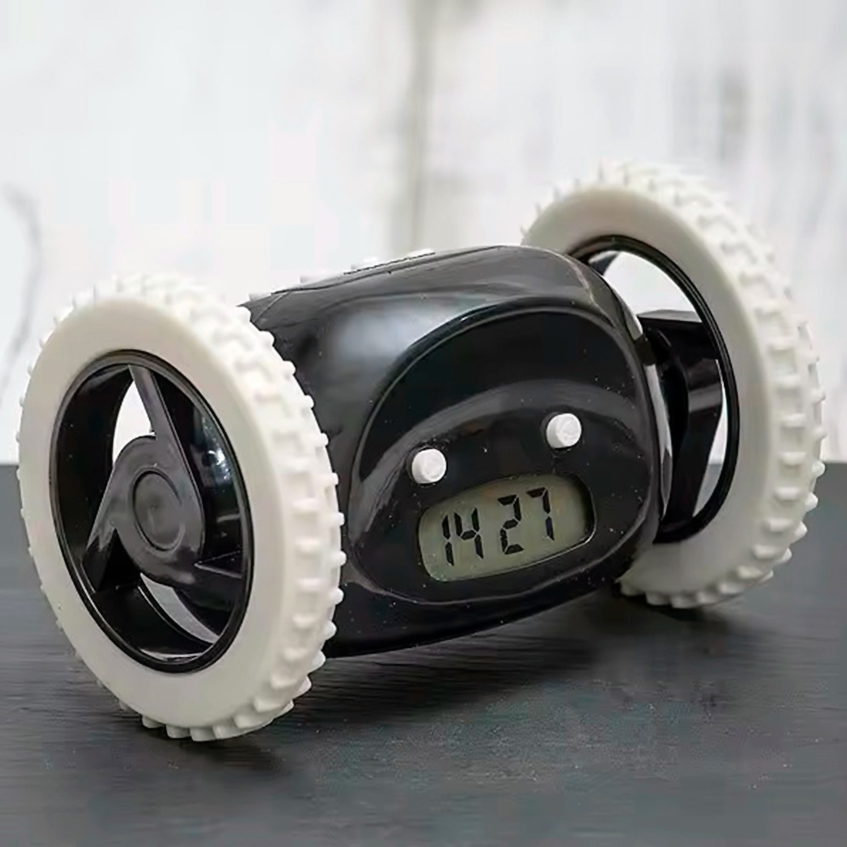 Un despertador negro con ruedas blancas muestra 14:27. El reloj está sobre una superficie oscura, con un fondo claro.