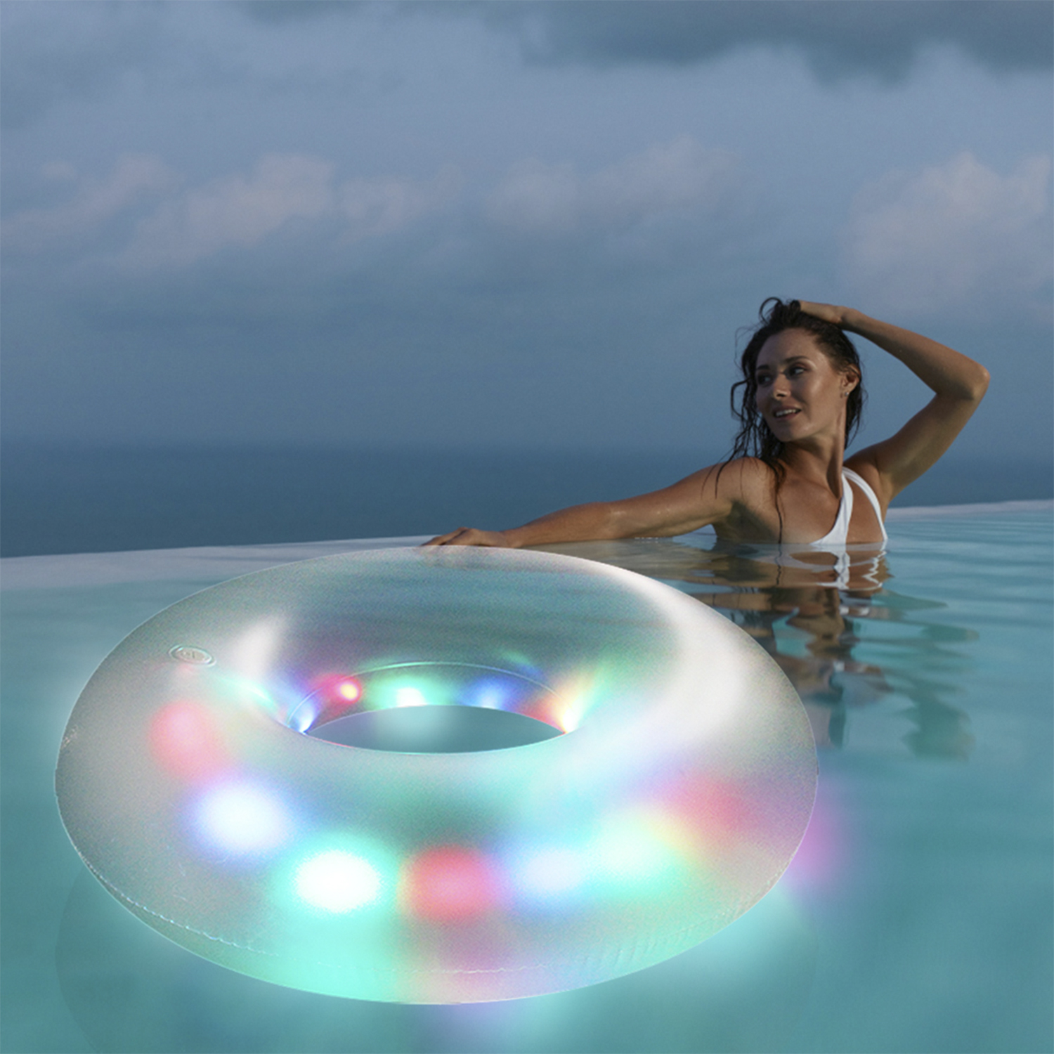 Donna in piscina con anello illuminato. Indossa un costume da bagno bianco. La piscina ha acqua limpida, sullo sfondo mare e cielo.