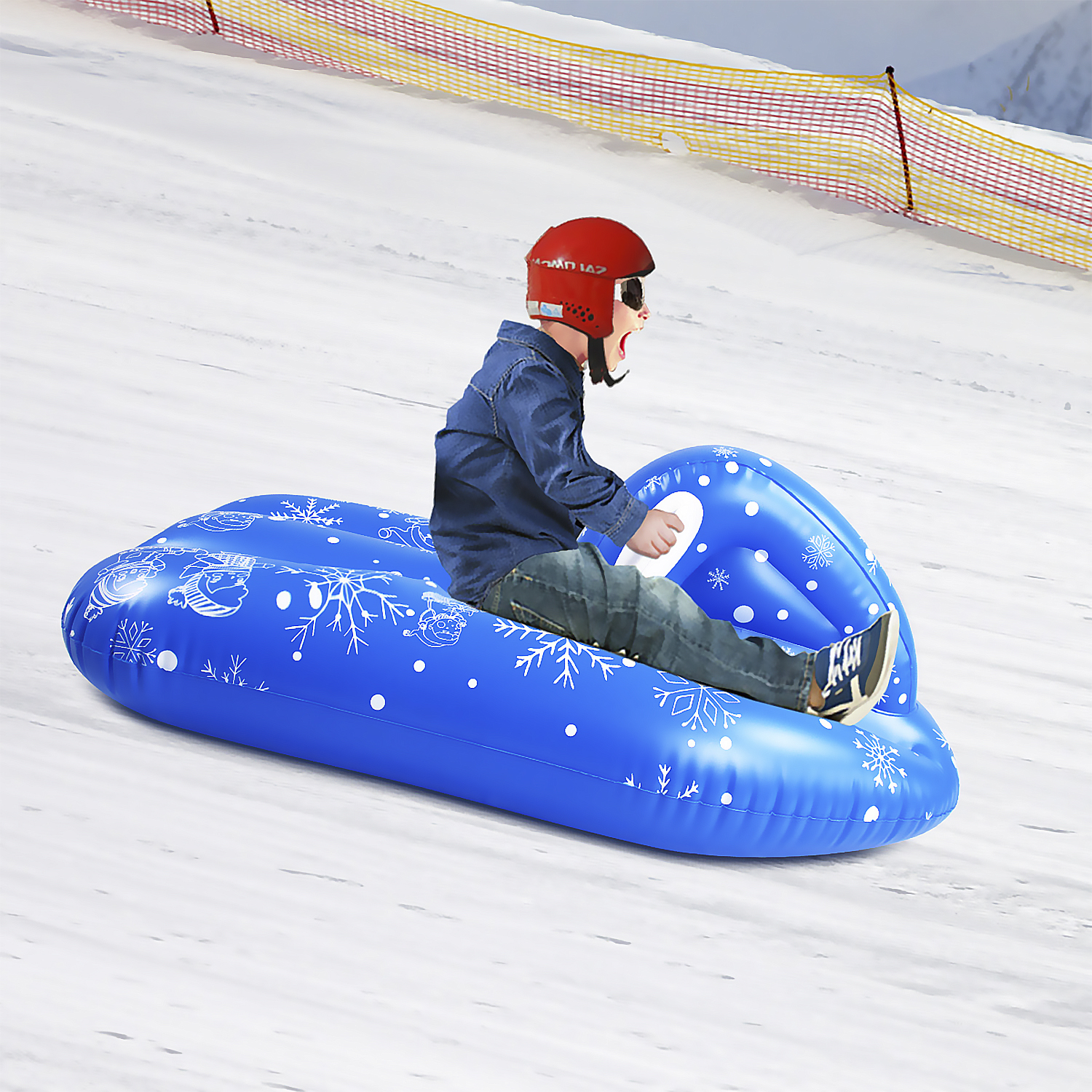 Un niño con casco rojo se desliza por una colina nevada en un trineo inflable azul con diseños de copos de nieve.