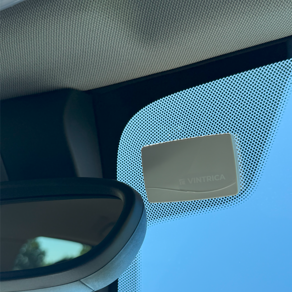 All'interno di un'auto, un dispositivo grigio con 'Vintrica' sul parabrezza, specchietto retrovisore e cielo blu visibile.