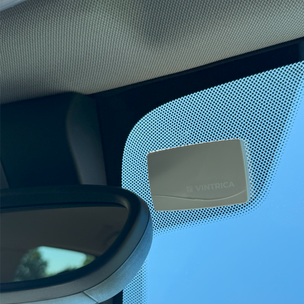 All'interno di un'auto, un dispositivo rettangolare con 'Vintrica' è montato sul parabrezza. Uno specchietto retrovisore è in basso a sinistra, cielo blu visibile.