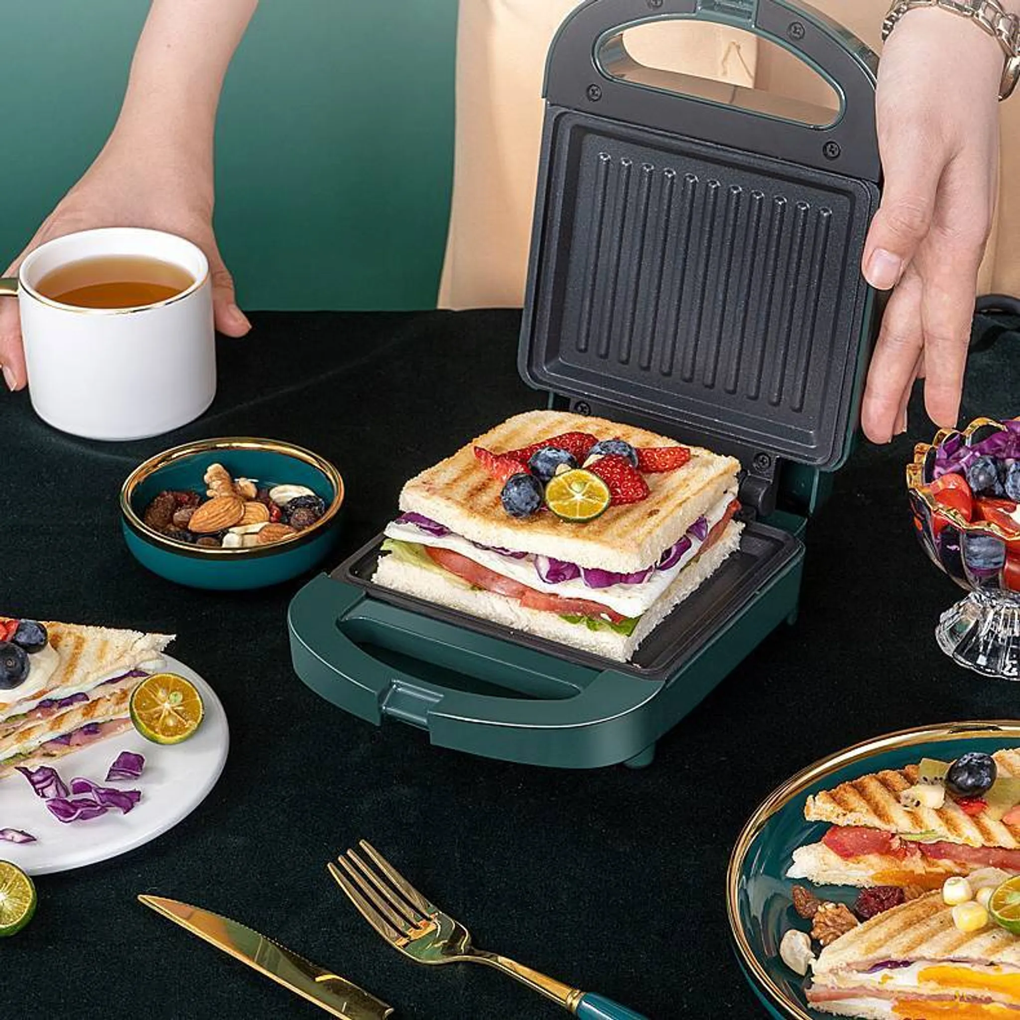 Sandwich in einem Sandwichmaker. Beeren oben drauf. Schalen mit Nüssen und Früchten. Eine Tasse Tee. Grüne und goldene Akzente.