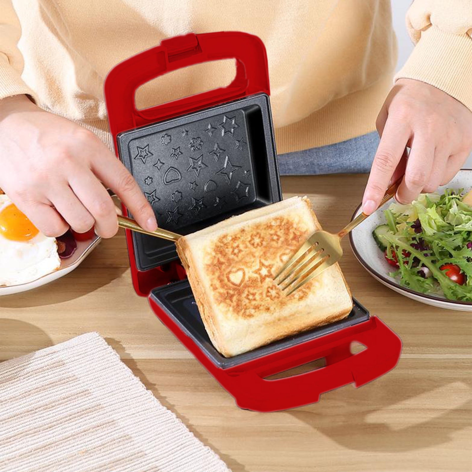 Ein roter Sandwichmaker mit einem getoasteten Sandwich. Hände halten Gabeln, und Teller mit Essen sind in der Nähe.
