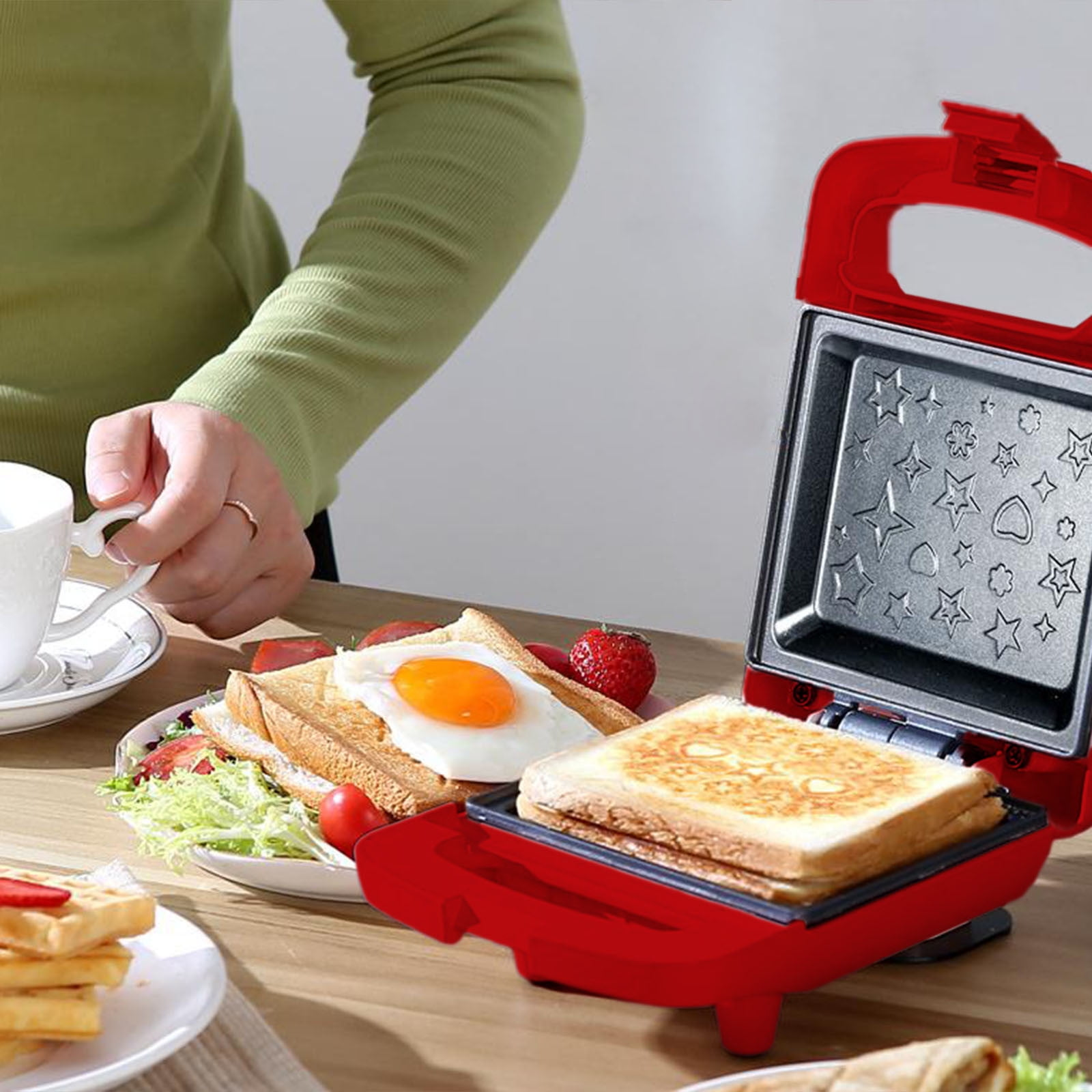 Eine Person bereitet das Frühstück zu. Ein roter Sandwichmaker ist geöffnet, mit einem Sandwich darin. Ein Teller hat ein Sandwich mit Spiegelei.