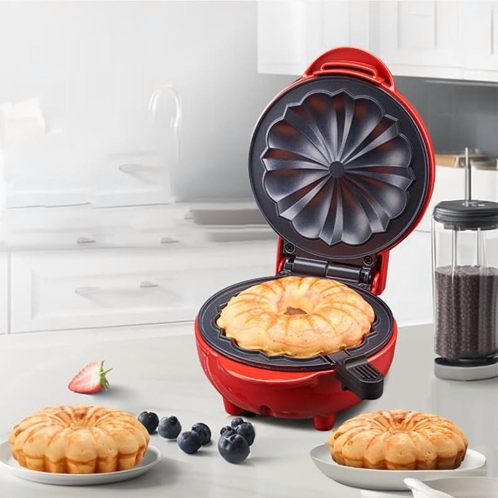 Ein roter Donut-Maker mit einem gebackenen Donut. Zwei Donuts auf Tellern, Blaubeeren und eine Erdbeerhälfte sind ebenfalls vorhanden.