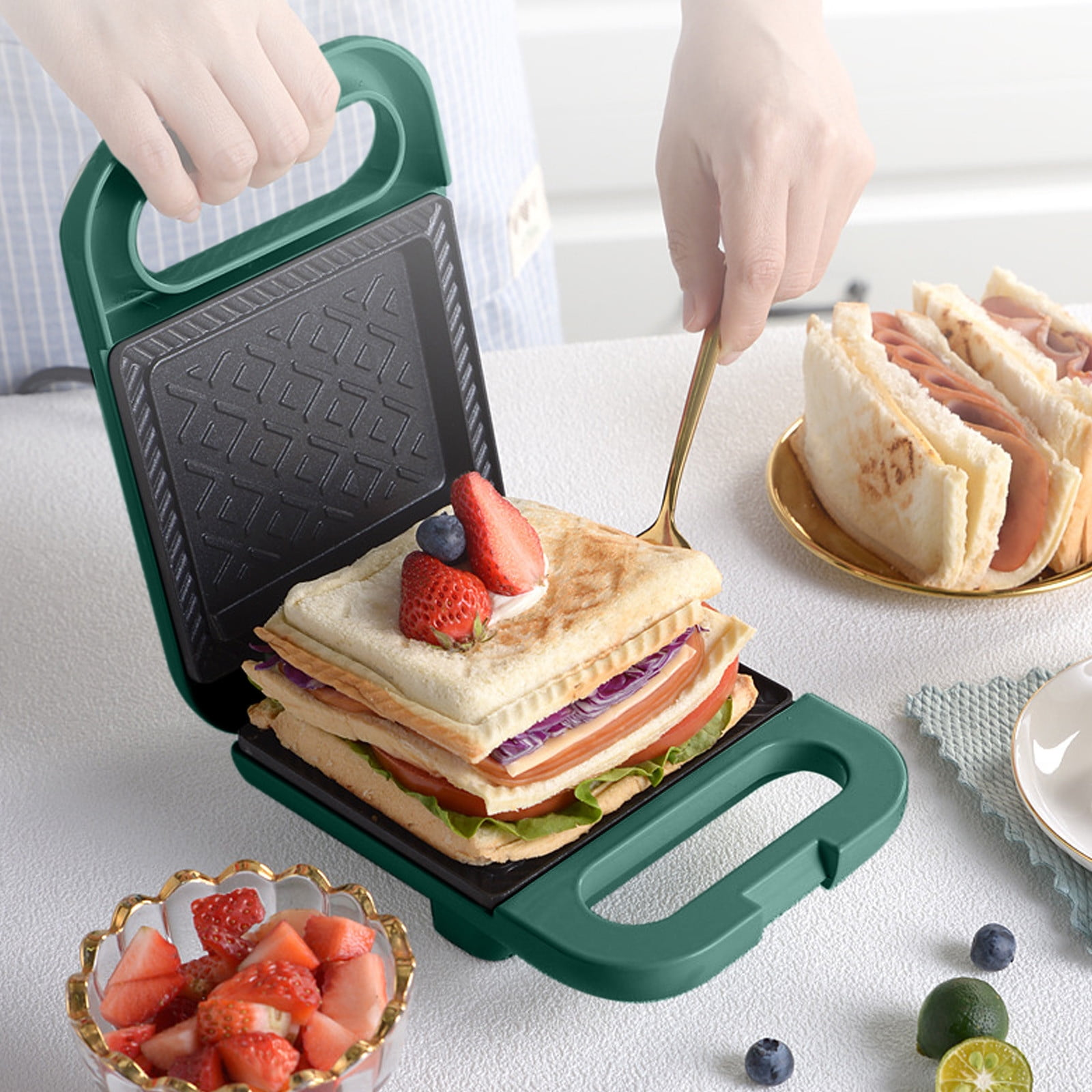 Ein Sandwichmaker mit einem Sandwich darin, garniert mit Beeren. Andere Sandwiches und eine Schüssel mit Erdbeeren sind in der Nähe.