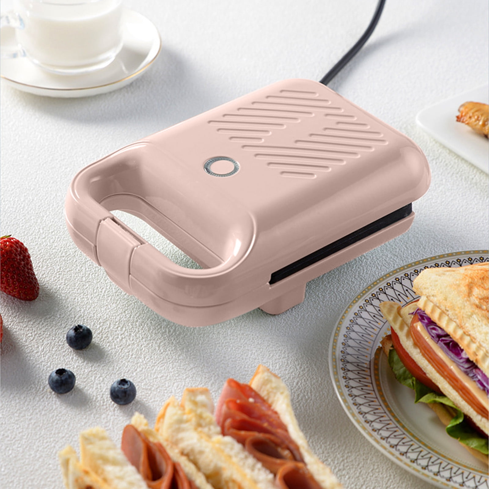 Ein rosa Sandwichmaker steht auf einer weißen Oberfläche. Sandwiches, Obst und ein Getränk sind in der Nähe.