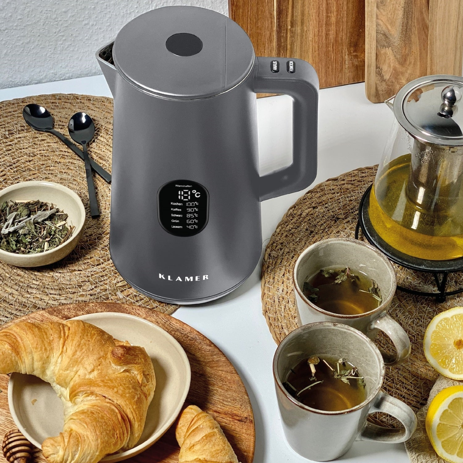 Ein grauer Wasserkocher mit Digitalanzeige, zwei Tassen Tee, ein Croissant, eine Teekanne und Zitronenscheiben auf einem Tisch.