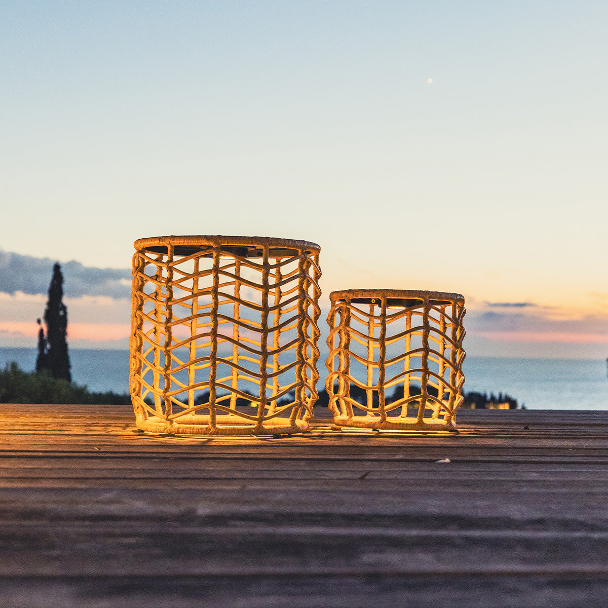 Zwei gewebte Laternen stehen auf einem Holzdeck in der Dämmerung. Der Himmel ist ein Gradient aus Orange und Blau. Das Meer und Bäume sind sichtbar.