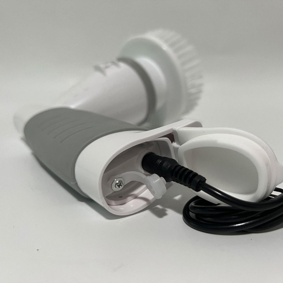 Eine weiße und graue elektrische Gesichtsbürste mit einem schwarzen Netzkabel liegt auf einer weißen Oberfläche. Der Bürstenkopf ist sichtbar.