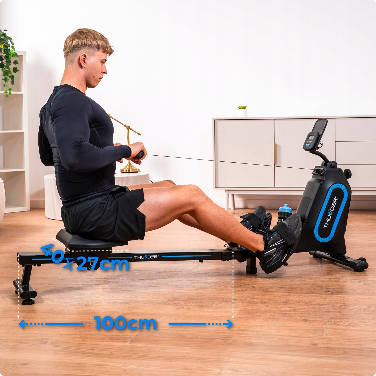 Mężczyzna korzysta z ergometru wioślarskiego. Ma na sobie czarny strój sportowy. Maszyna jest czarno-niebieska. Tło to pokój z białymi ścianami.