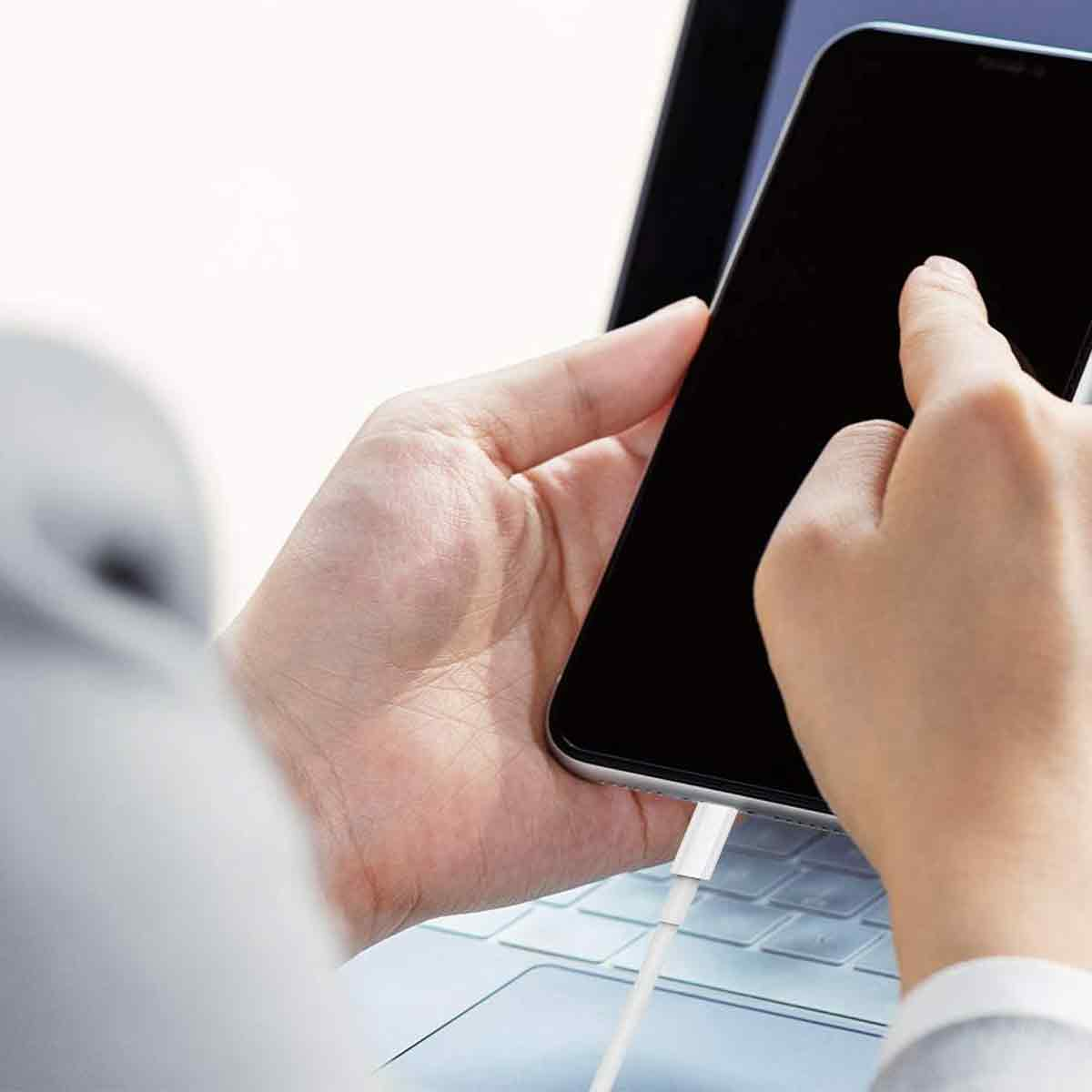 Eine Hand hält ein schwarzes Smartphone, das mit einem weißen Ladekabel verbunden ist. Ein Finger berührt den Bildschirm. Ein Laptop ist im Hintergrund.