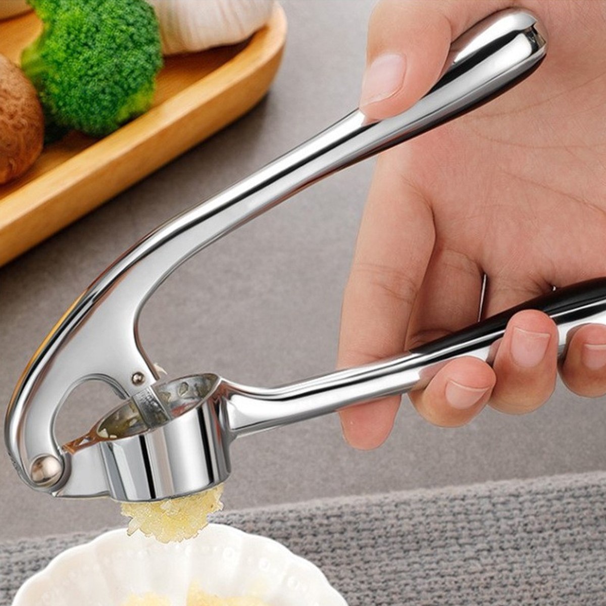 Eine Hand drückt eine Knoblauchpresse und extrudiert gehackten Knoblauch auf einen weißen Teller. Brokkoli und Knoblauch sind im Hintergrund.