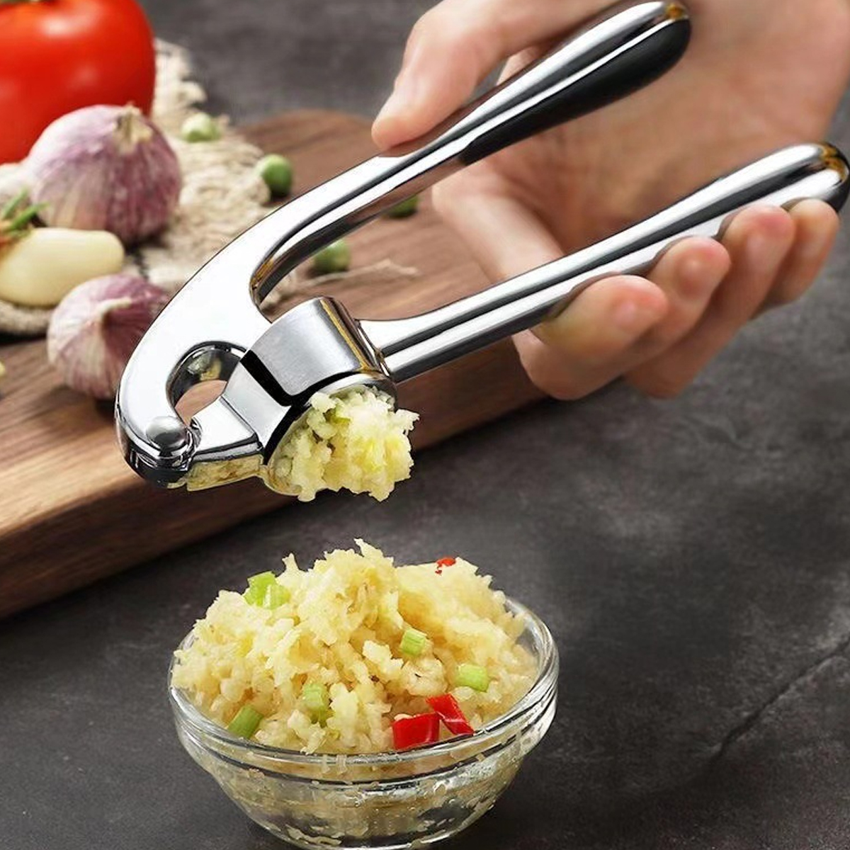 Eine Hand drückt eine Knoblauchpresse und extrudiert gehackten Knoblauch. Eine Schüssel enthält den gehackten Knoblauch. Knoblauchzehen und Tomate sind in der Nähe.