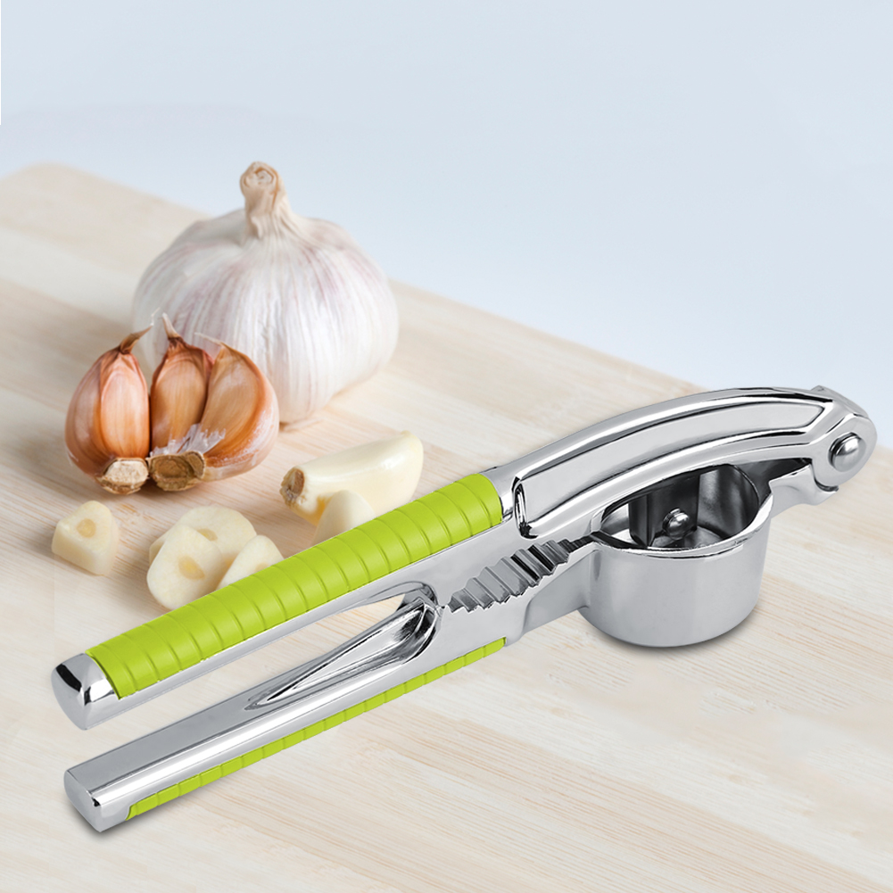 Eine Knoblauchpresse mit grünem Griff liegt auf einem Holzbrett mit Knoblauchzehen und einer ganzen Knoblauchzehe.