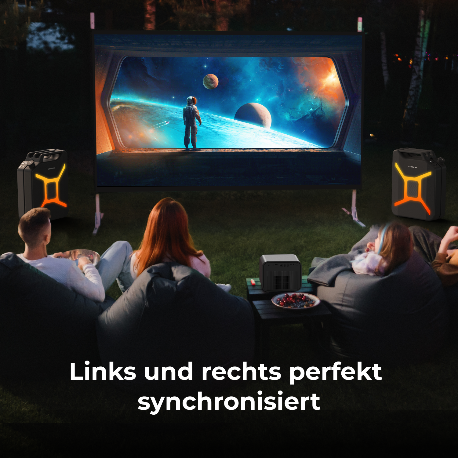 Leute schauen sich im Freien eine Weltraumszene an, die auf eine Leinwand projiziert wird. Lautsprecher mit orangem Licht flankieren die Leinwand.