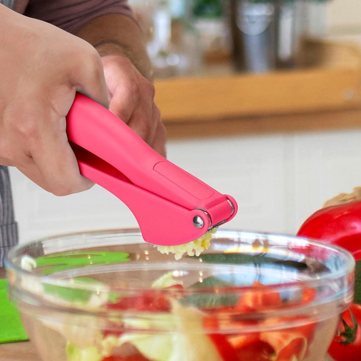 Eine Person benutzt eine pinkfarbene Knoblauchpresse über einer Glasschüssel mit Gemüse. Der Knoblauch wird gepresst. Roter Pfeffer ist ebenfalls sichtbar.