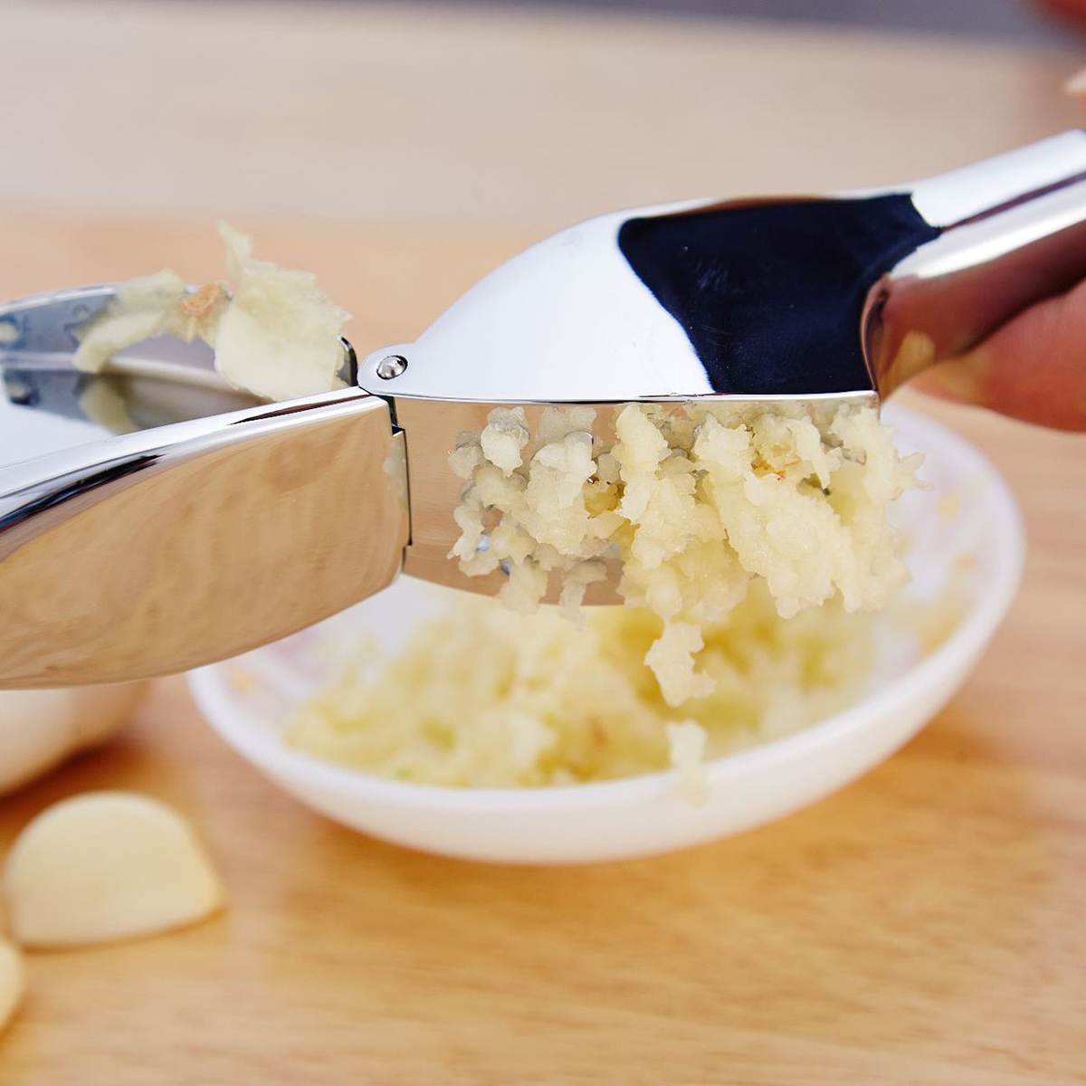 Eine Knoblauchpresse zerdrückt Knoblauch. Zerdrückter Knoblauch fällt in eine weiße Schüssel. Einige Knoblauchzehen liegen auf Holz.