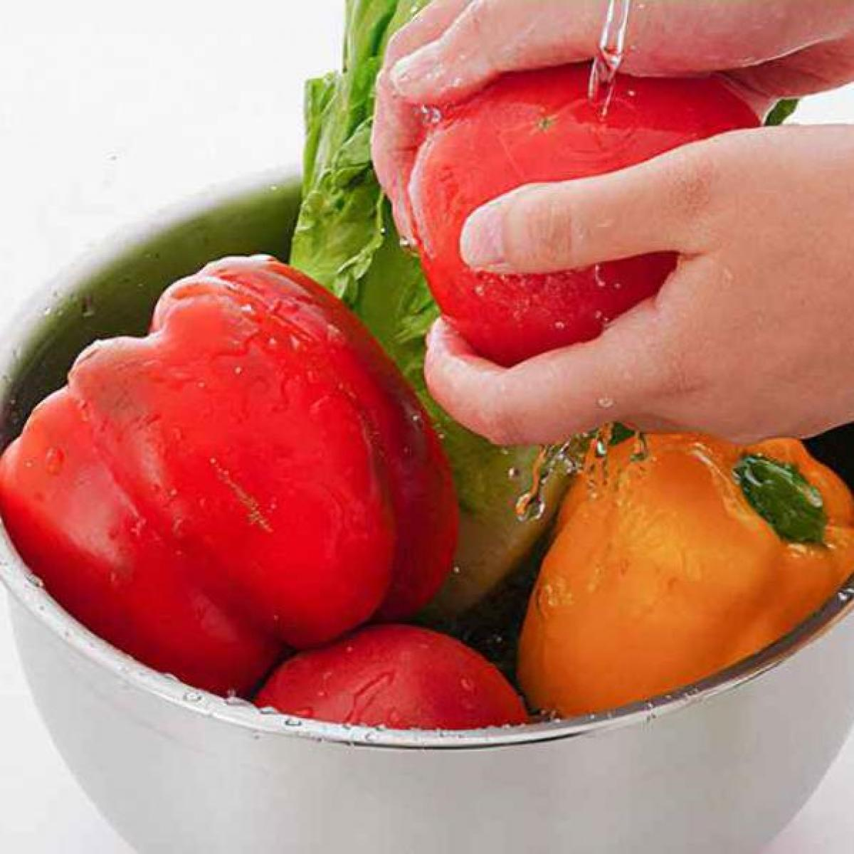 Hände waschen eine rote Tomate unter fließendem Wasser in einer Metallschüssel. Paprika und Salat sind ebenfalls in der Schüssel.