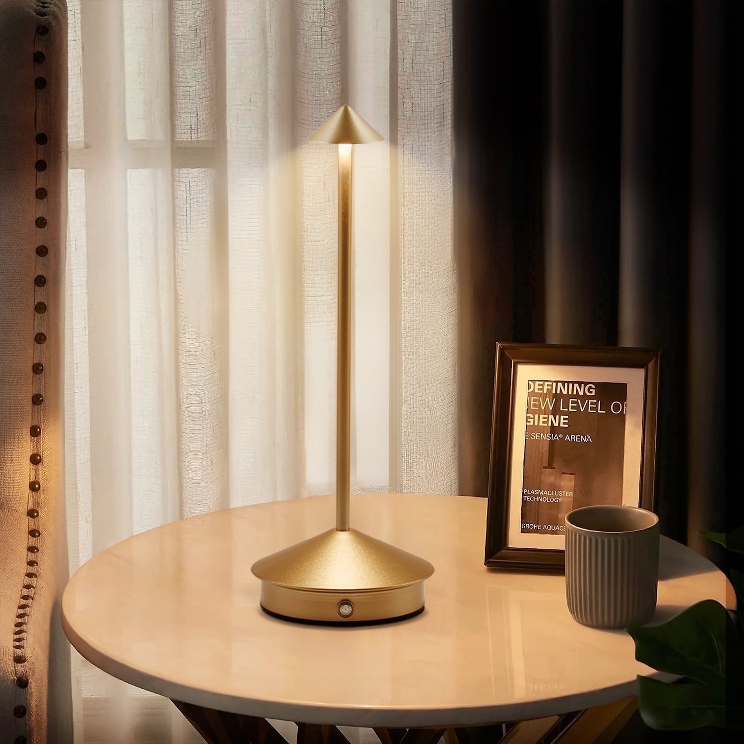 Eine goldene Lampe auf einem Tisch. Dahinter Vorhänge, ein Bilderrahmen und eine Tasse. Die Lampe ist beleuchtet.