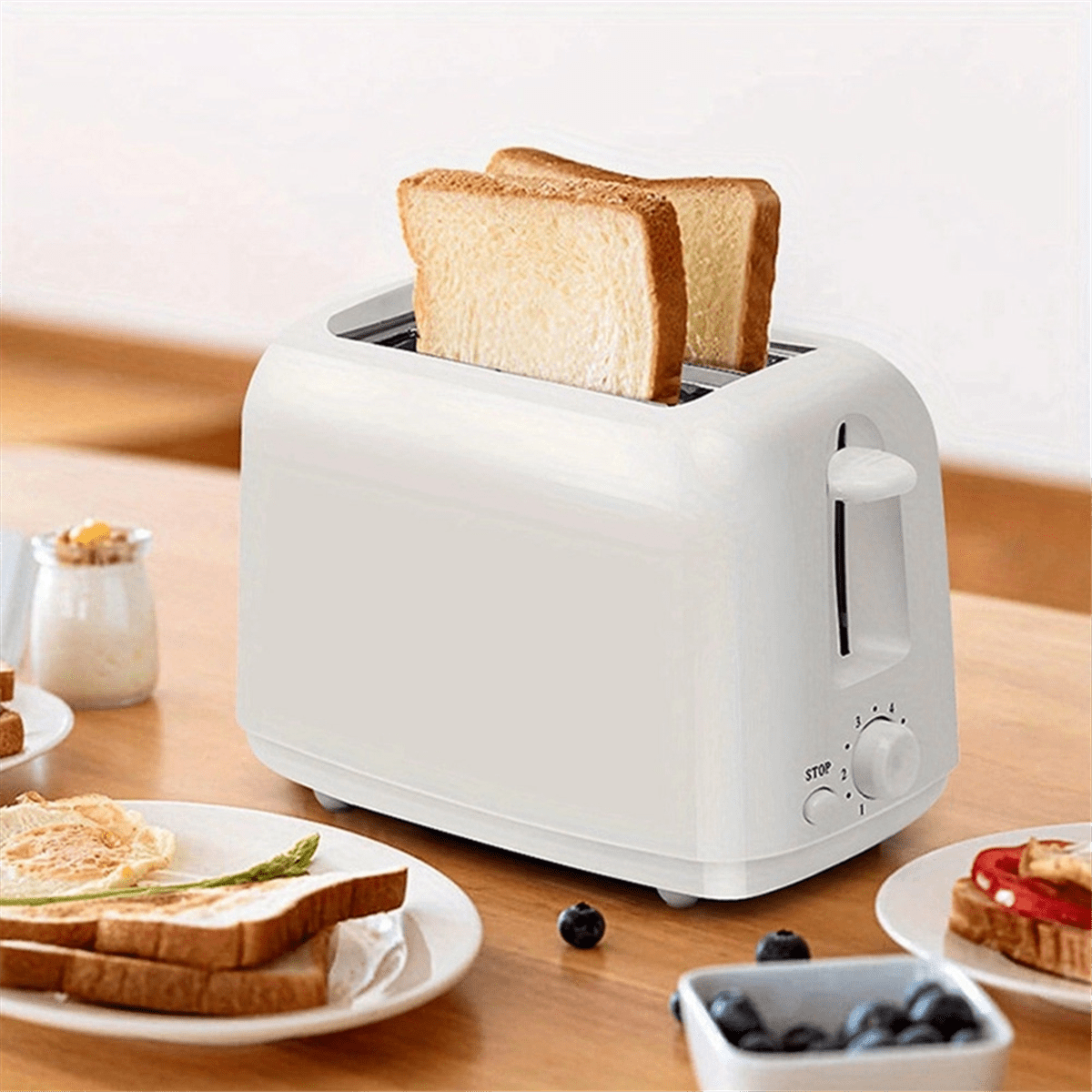 Ein weißer Toaster mit zwei Toastscheiben steht auf einem Holztisch. Teller mit Essen und Blaubeeren sind im Vordergrund.