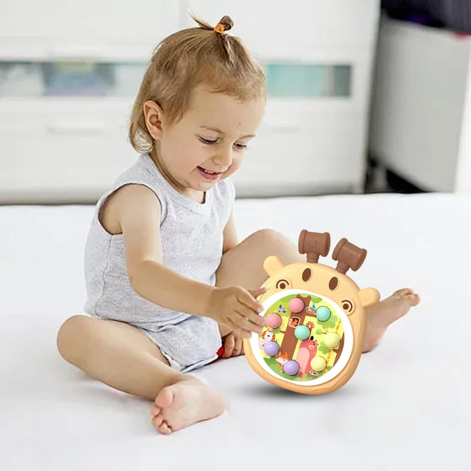 Ein lächelndes Kleinkind spielt mit einem giraffenförmigen Spielzeug. Das Spielzeug hat bunte Bälle, einen Hammer und Tierbilder. Die Szene ist drinnen.
