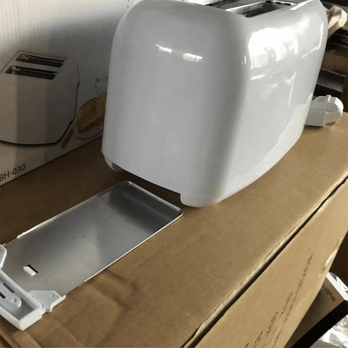 Ein weißer Toaster steht auf einem Karton. Davor befindet sich ein silbernes Krümelblech. Ein weiterer Toaster ist im Hintergrund teilweise sichtbar.