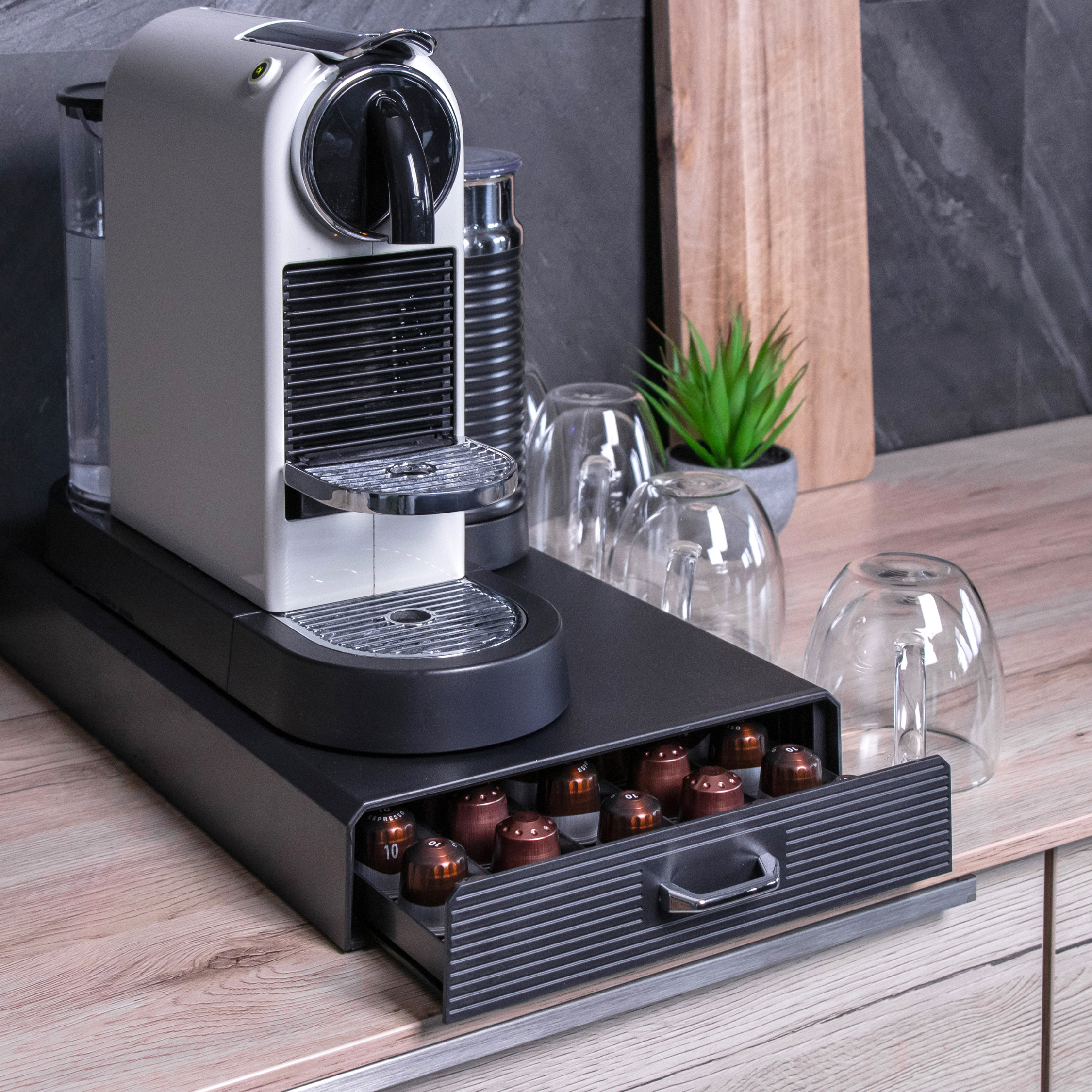 Eine weiß-schwarze Kaffeemaschine mit Kapseln in einer Schublade, in der Nähe von Gläsern und einer kleinen Pflanze.