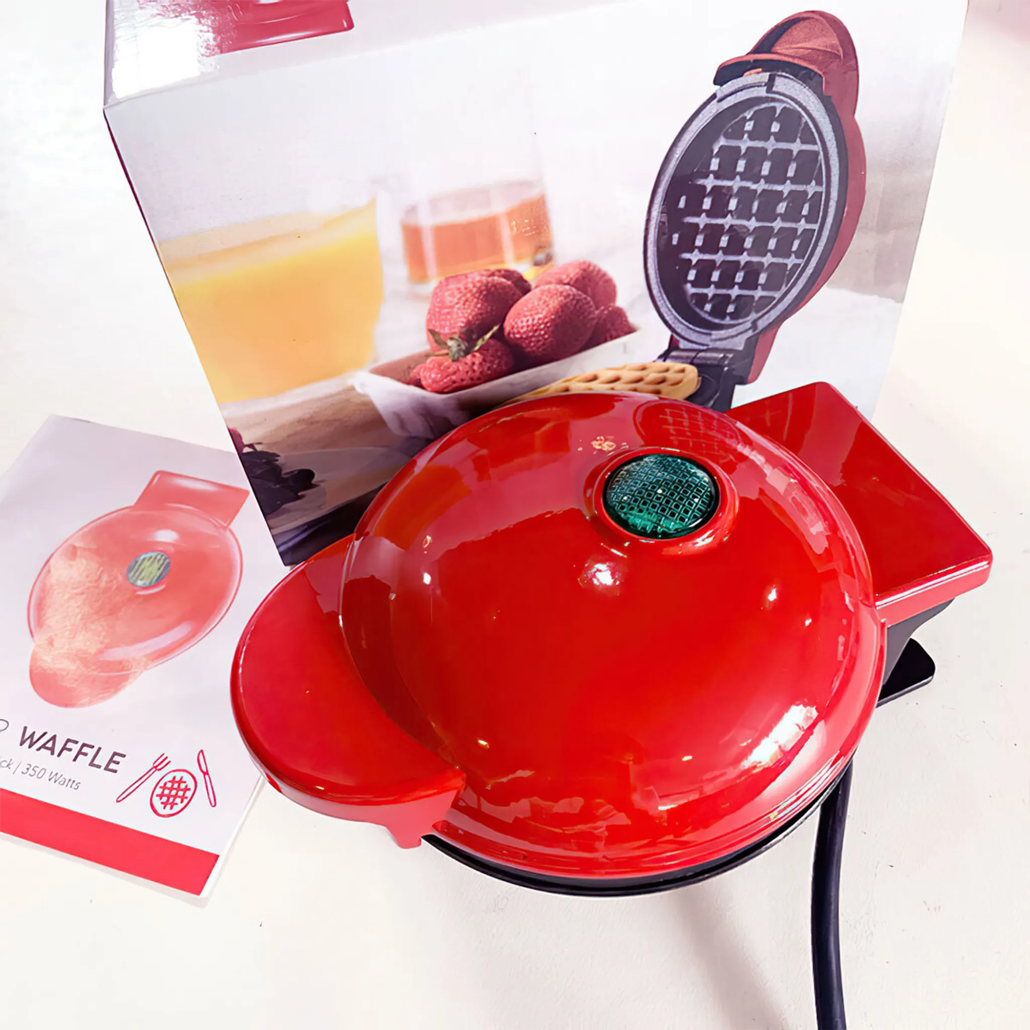 Ein roter Waffeleisen mit grünem Licht. Eine Schachtel zeigt Waffeln, Erdbeeren und Saft. Auf der linken Seite befindet sich ein Faltblatt.