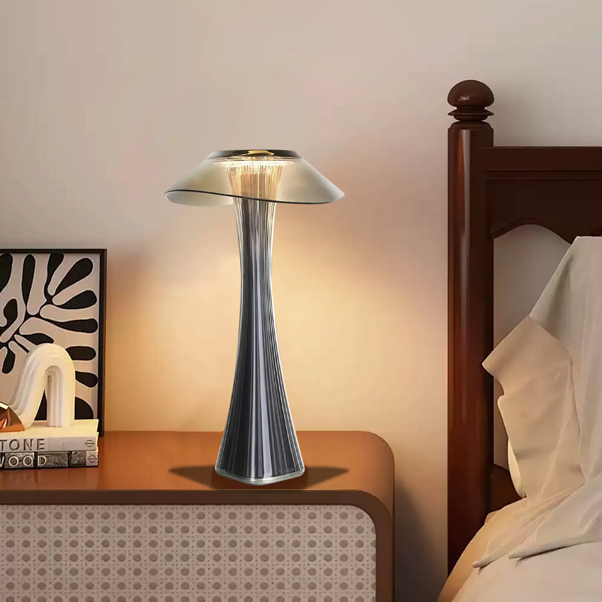Eine moderne Lampe auf einem Nachttisch neben einem Bett. Ein gerahmtes Kunstwerk und Bücher stehen ebenfalls auf dem Ständer.