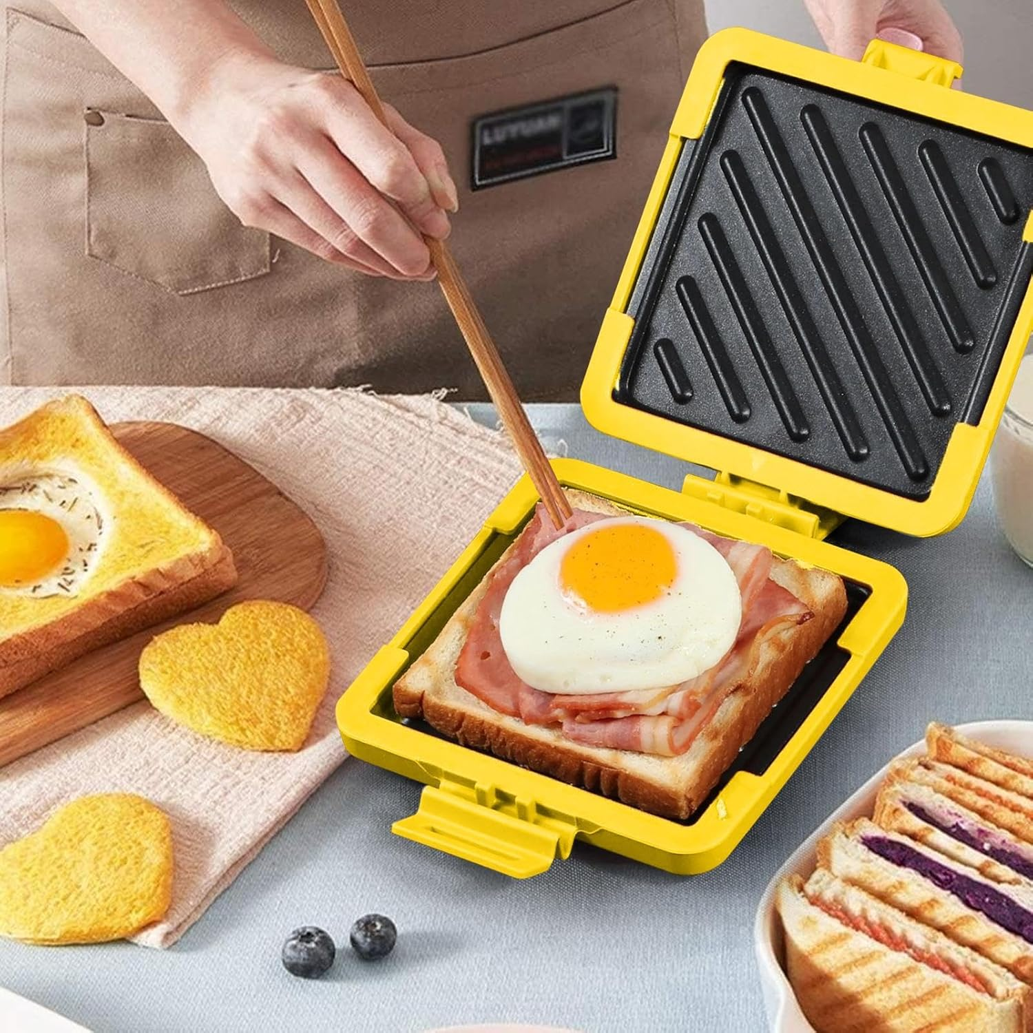 Eine Person benutzt einen gelben Sandwichmaker, um Ei und Speck auf Brot zu kochen. Anderes Brot mit Ei und herzförmiges Brot liegen daneben.
