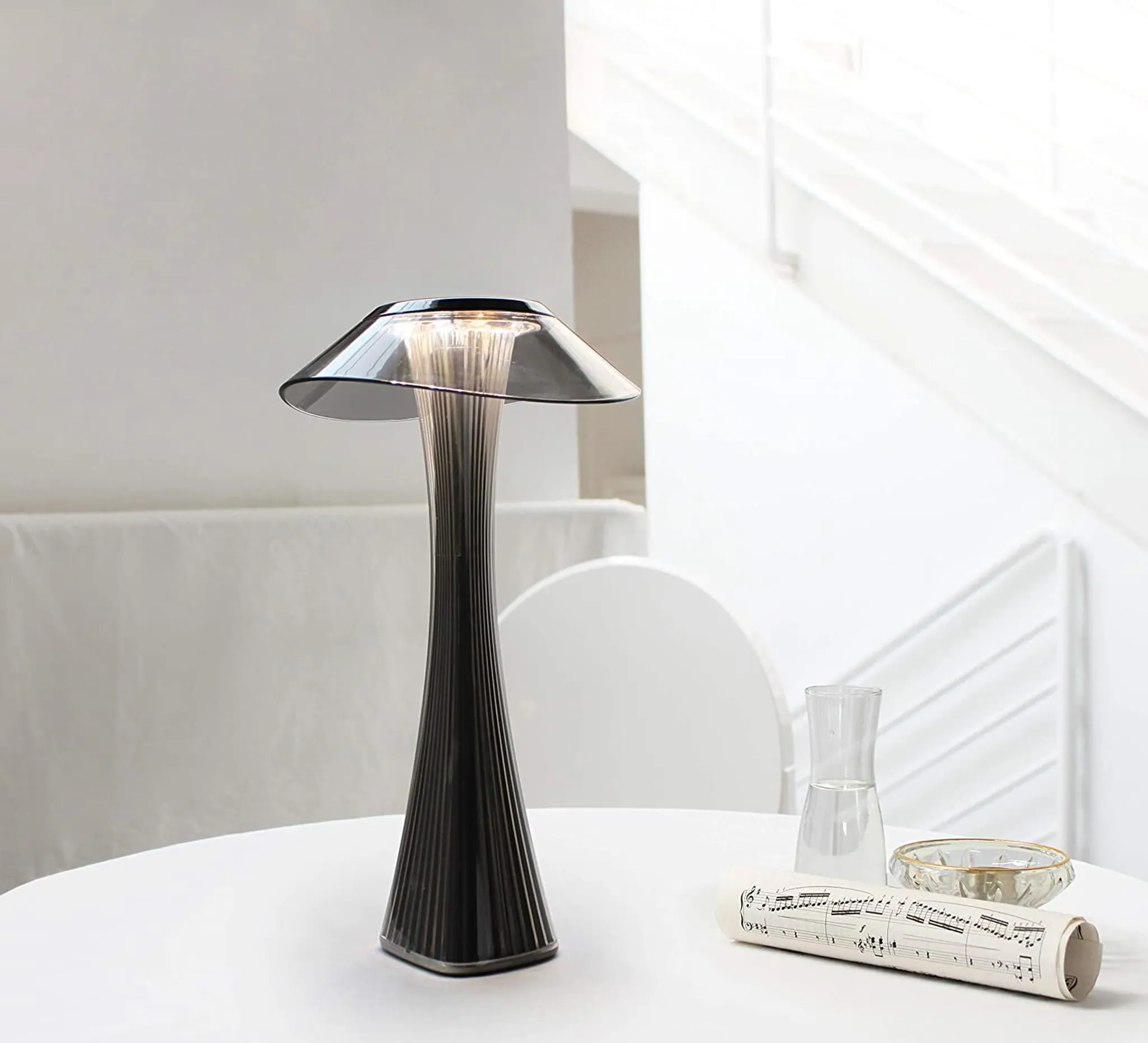 Eine moderne Lampe mit schwarzem Fuß und grauem Schirm steht auf einem weißen Tisch. Ein Glas Wasser und ein Notenblatt liegen auf dem Tisch.