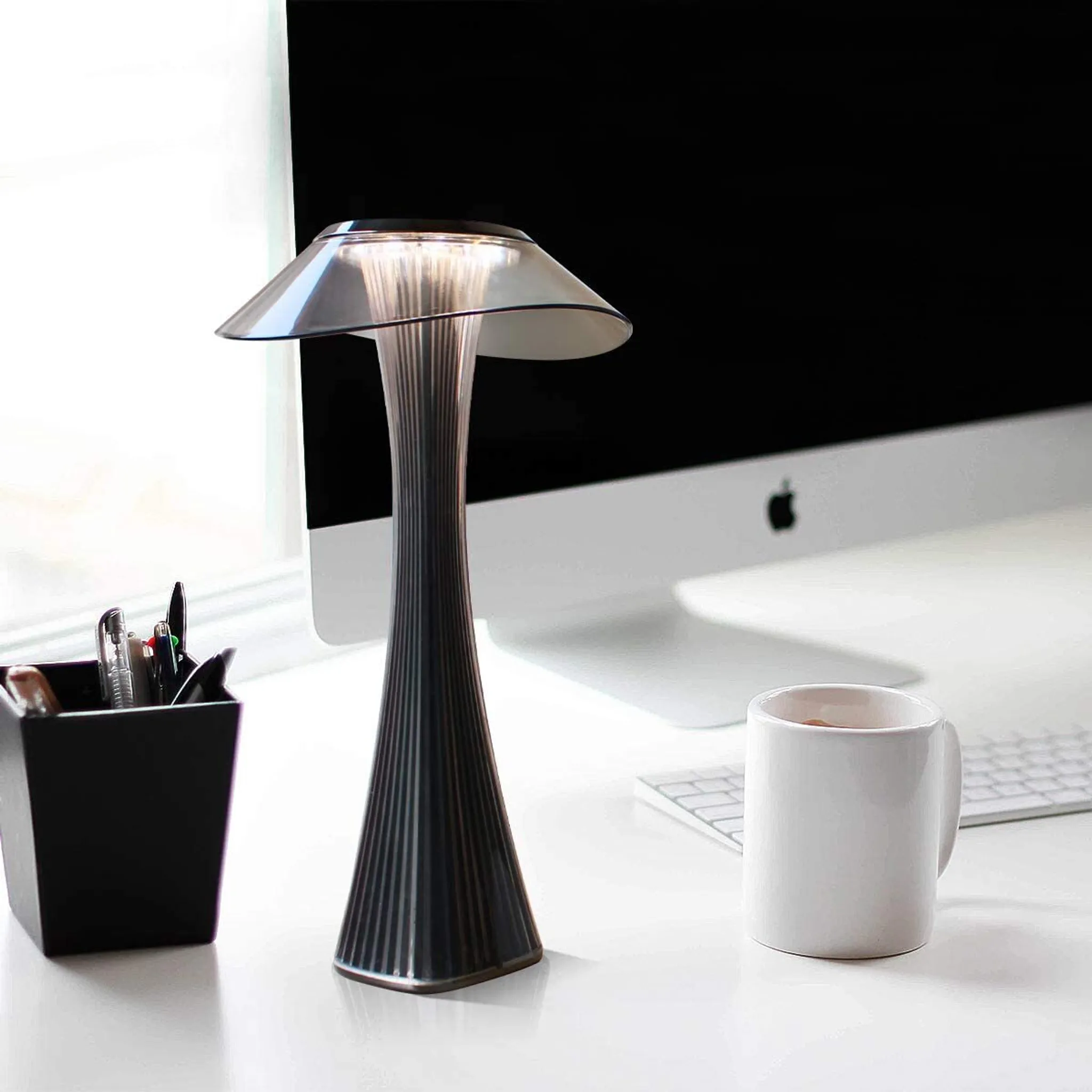 Schreibtisch mit Lampe, Tasse, Computer und Stifthalter. Lampe ist an. Computerbildschirm ist dunkel. Weißer Schreibtisch.