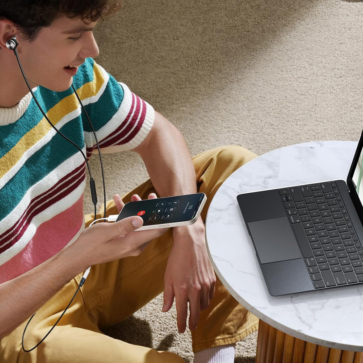 Ein Mann mit Kopfhörern hält ein Telefon, auf einem Tisch steht ein Laptop. Das Telefon zeigt einen Anrufbildschirm.