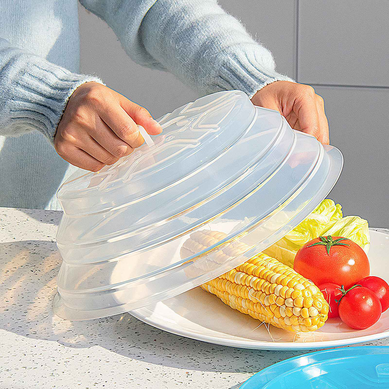 Hände heben einen transparenten Essensbehälter an. Auf einem Teller Mais, Tomaten und Salat. Hintergrund: helle Wand und Theke.