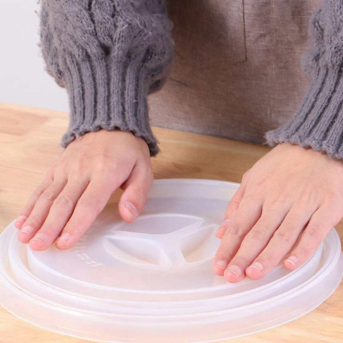 Hände legen einen weißen Kunststoffdeckel auf einen Behälter. Die Person trägt einen grauen Pullover und eine braune Schürze.