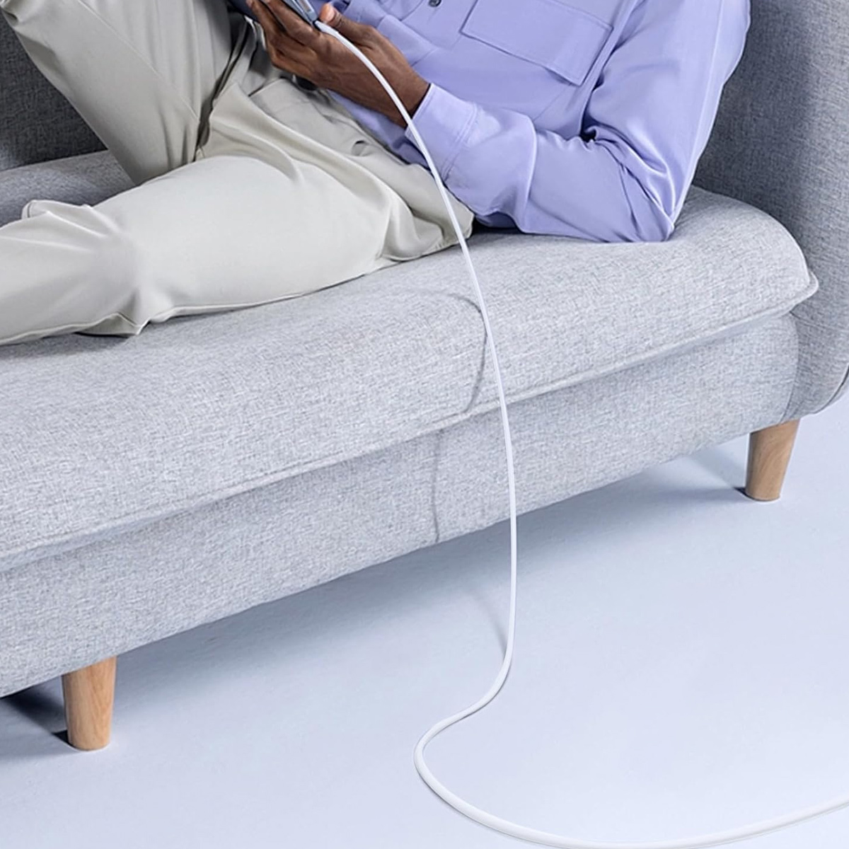 Person auf einem grauen Sofa, trägt ein lila Hemd, hält ein Telefon, verbunden mit einem weißen Kabel. Der Hintergrund ist hellblau.