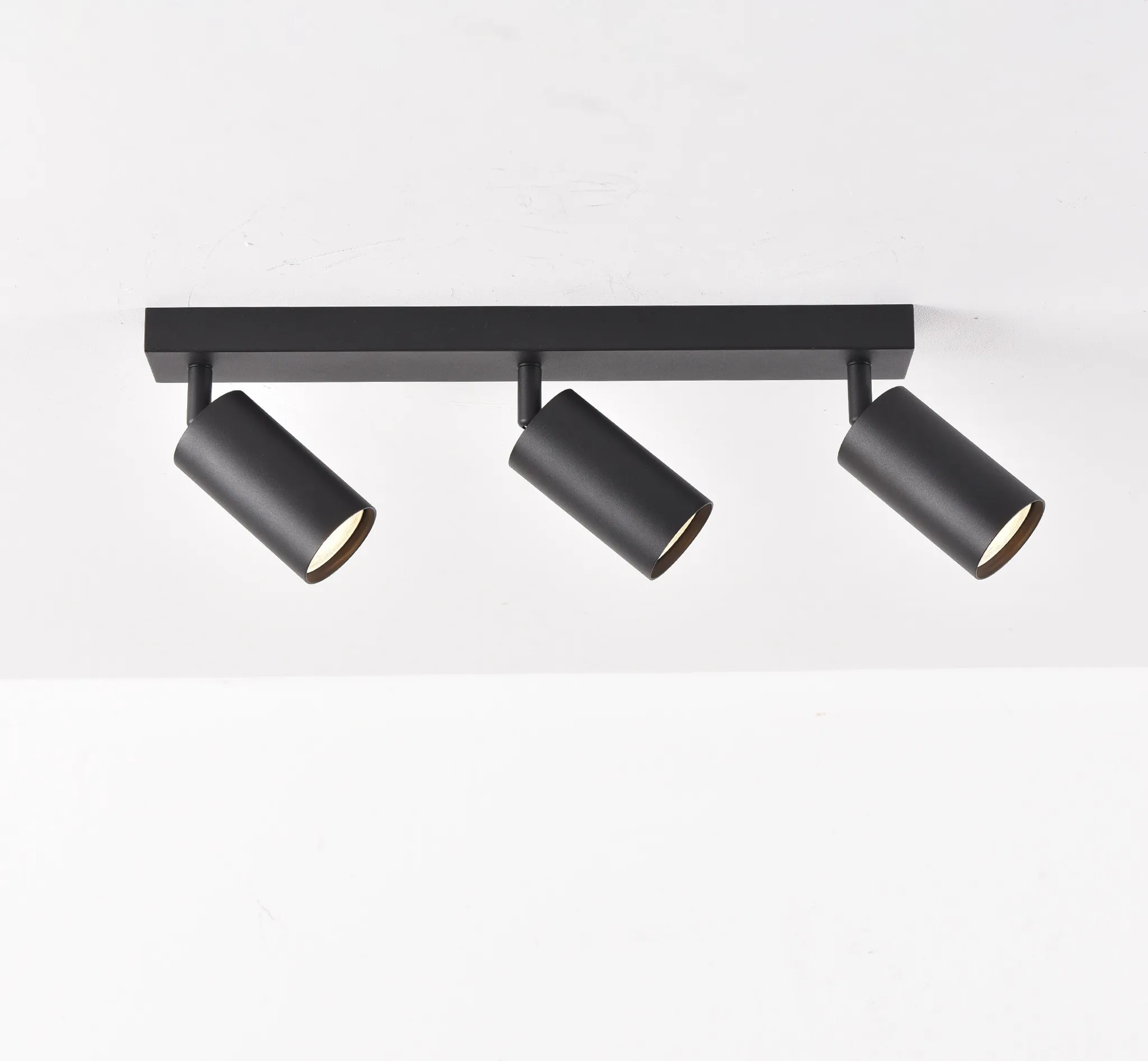 Schwarze Deckenleuchte mit drei verstellbaren Strahlern vor einer weißen Wand. Die Lichter sind an.