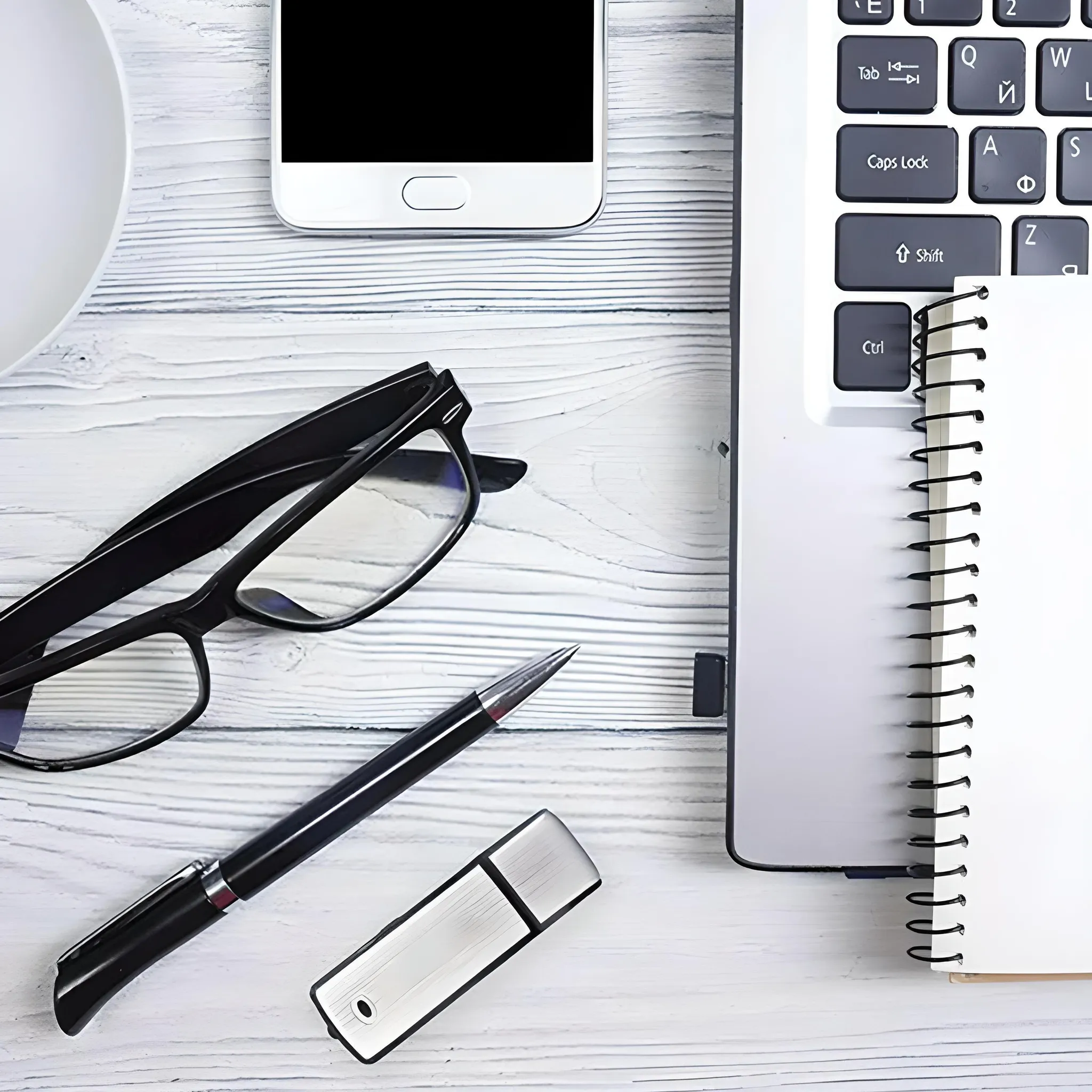 Schreibtisch-Setup: Telefon, Brille, Stift, USB, Laptop und Notizbuch auf weißer Holzoberfläche. Draufsicht.