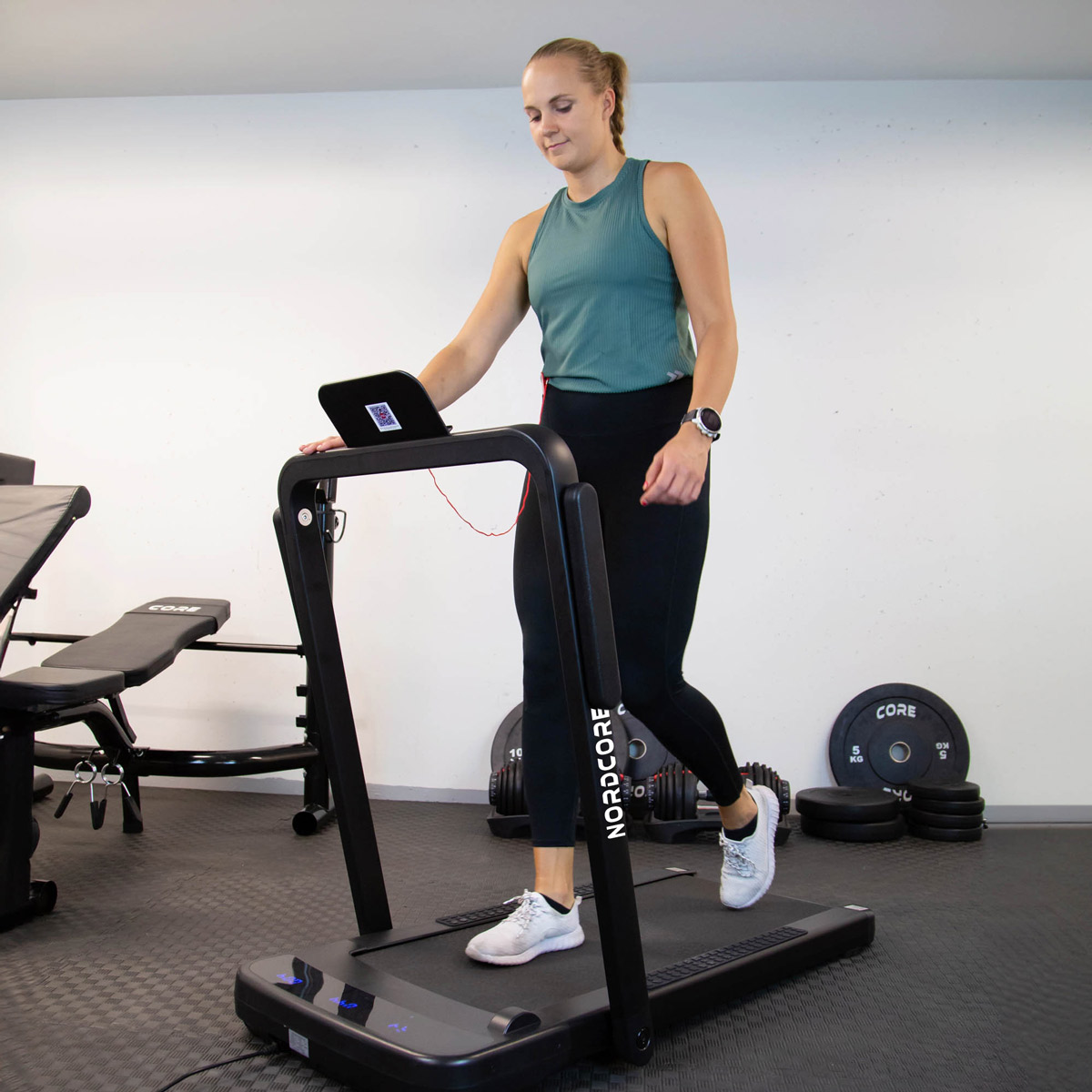 Kobieta ćwiczy na bieżni w siłowni. Ma na sobie zielony top, czarne legginsy i białe trampki. Sprzęt do ćwiczeń znajduje się w tle.