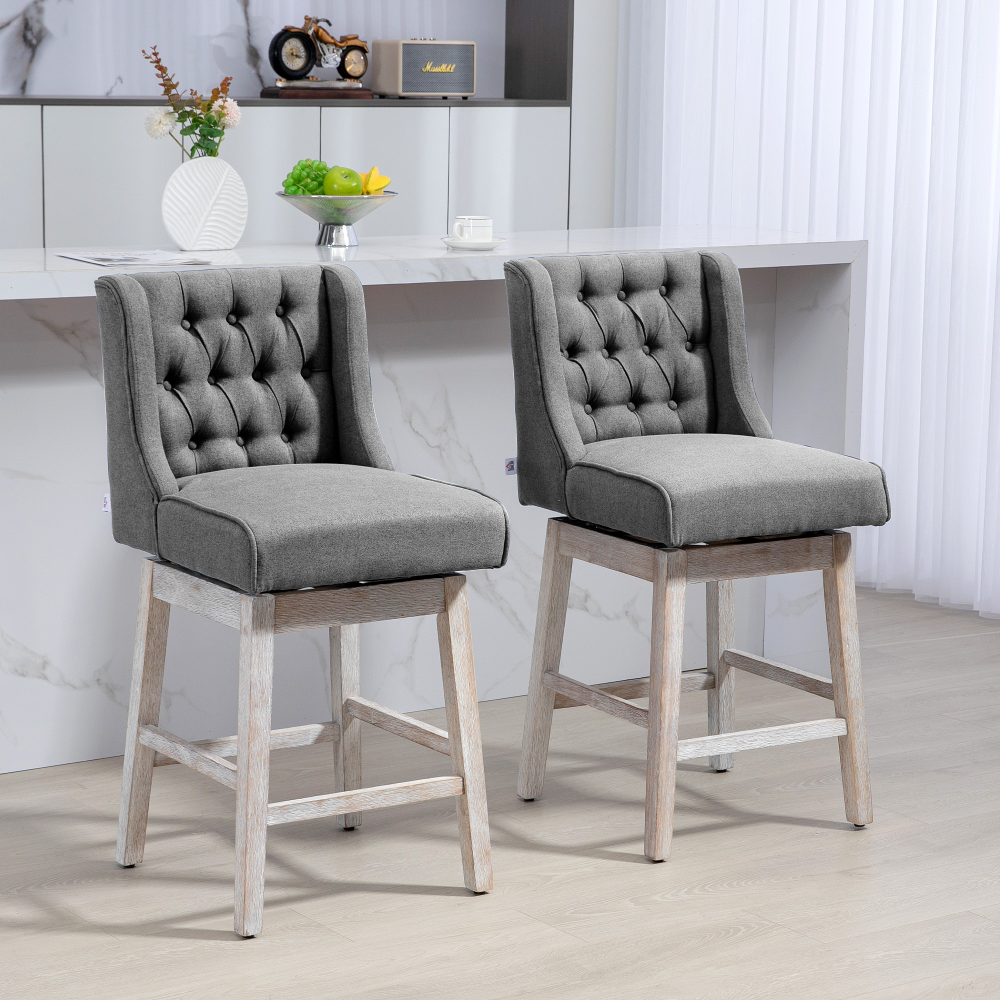 Dos taburetes de bar tapizados en gris con patas de madera blanca. Una encimera de mármol con fruta y un jarrón decorativo.