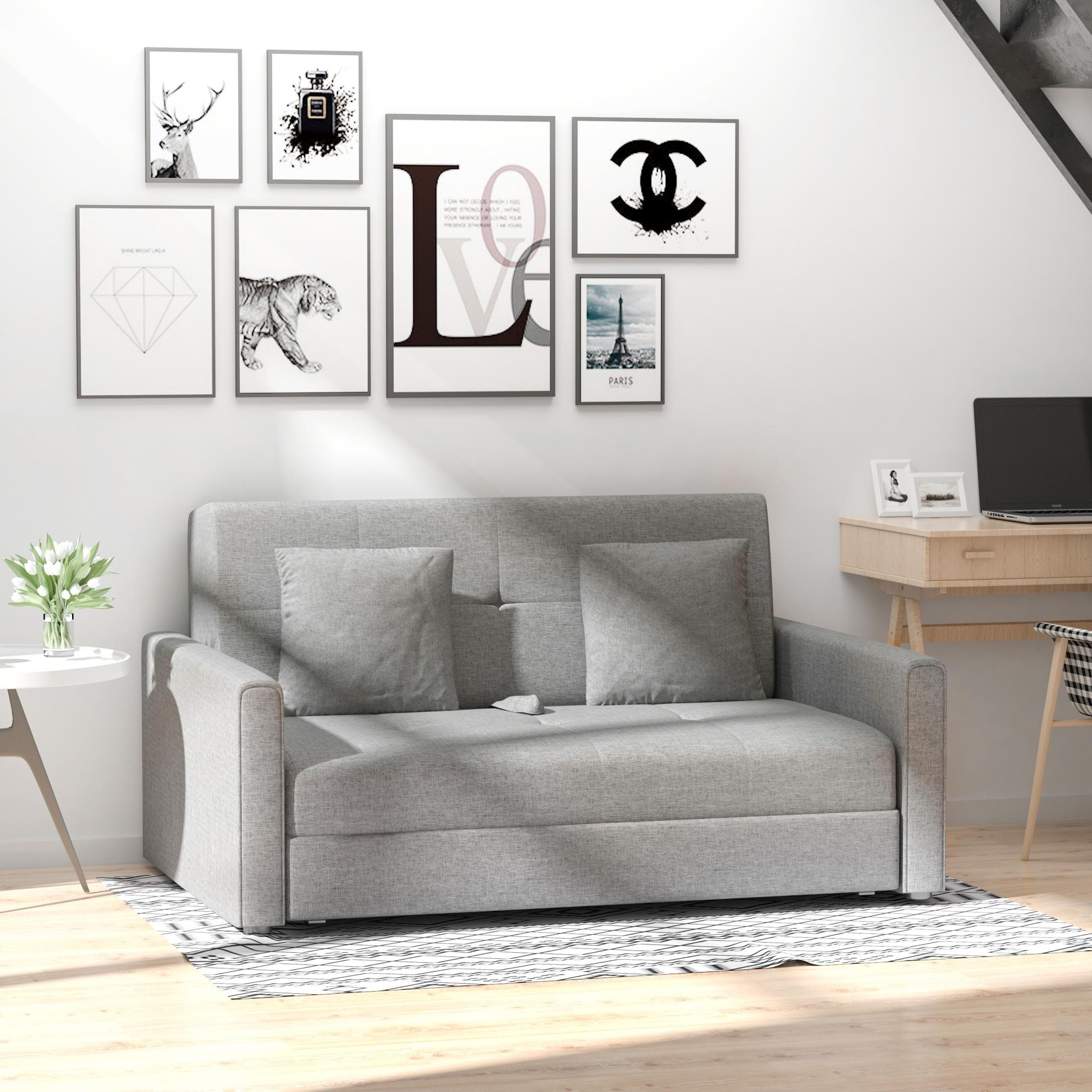 Un sofá gris con almohadas se encuentra debajo de arte enmarcado. Un escritorio con una laptop y una mesa con flores están cerca.